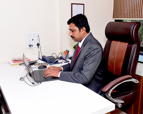 person writing in a notebook beside by an iPad, laptop, printed photos, spectacles, and a cup of coffee on a saucer