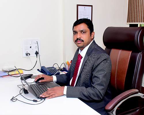 person writing in a notebook beside by an iPad, laptop, printed photos, spectacles, and a cup of coffee on a saucer